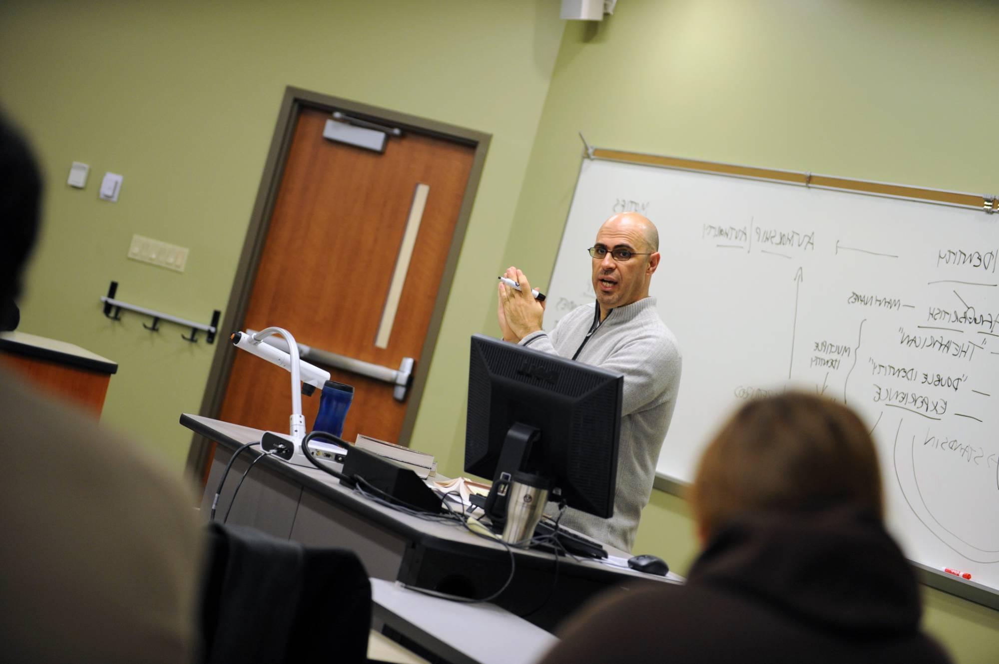 Male faculty member in the middle of teaching, standing near a white board with a white board marker in his hand as he explains a concept.